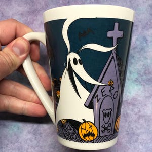 May include: White ceramic mug featuring Zero from The Nightmare Before Christmas. The design includes Zero, a tombstone with the word "ZERO", jack-o'-lanterns, and a bat against a dark teal background. The mug has a white handle.