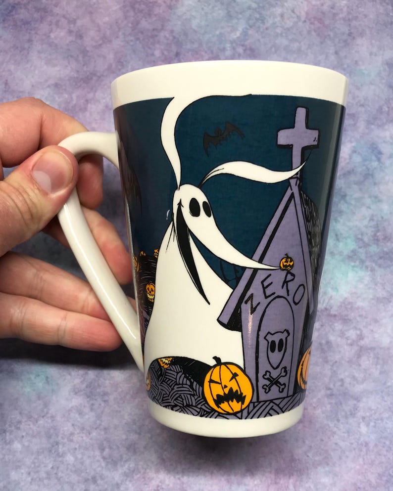 May include: White ceramic mug featuring Zero from The Nightmare Before Christmas. The design includes Zero, a tombstone with the word "ZERO", jack-o'-lanterns, and a bat against a dark teal background. The mug has a white handle.