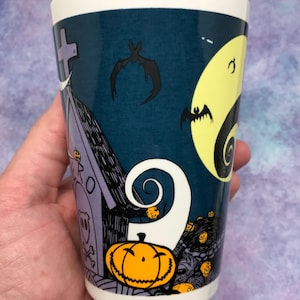May include: A white ceramic tumbler featuring a spooky Halloween design. The design includes a graveyard scene with a tombstone, bats, a full moon, and a grinning jack-o'-lantern. The tumbler has a dark blue background and a white rim.