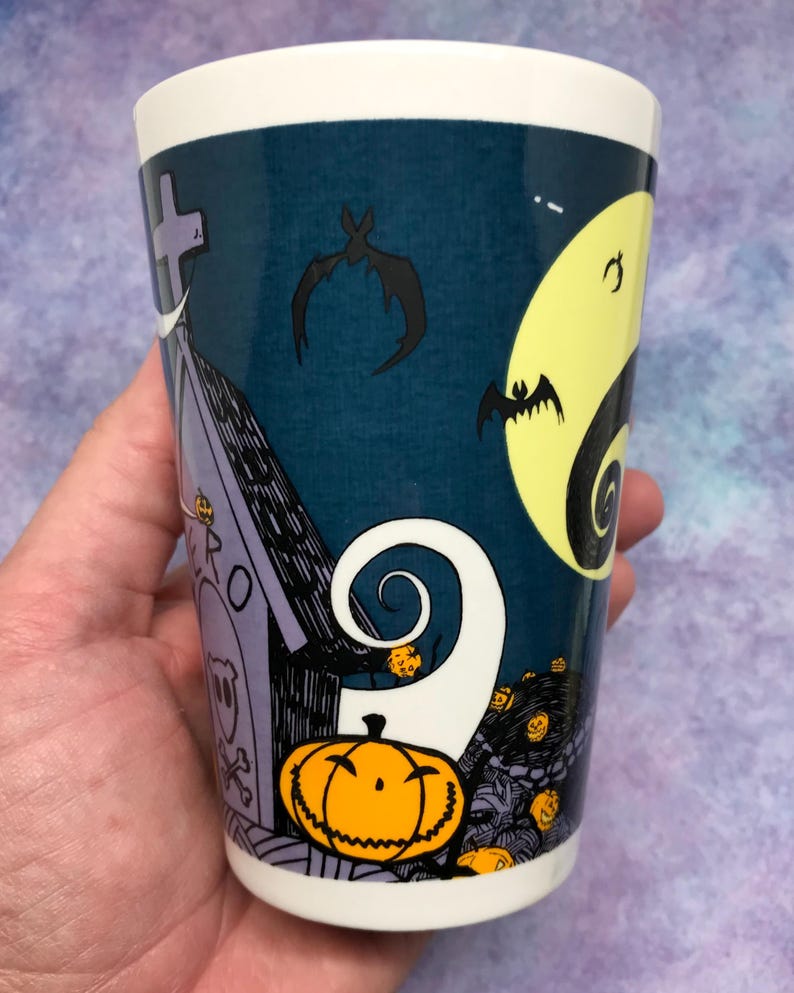 May include: A white ceramic tumbler featuring a spooky Halloween design. The design includes a graveyard scene with a tombstone, bats, a full moon, and a grinning jack-o'-lantern. The tumbler has a dark blue background and a white rim.
