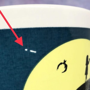 May include: A ceramic mug with a dark blue background and a yellow moon design. The mug features black silhouettes of a bat, a figure, and a tree. The top of the mug is white.