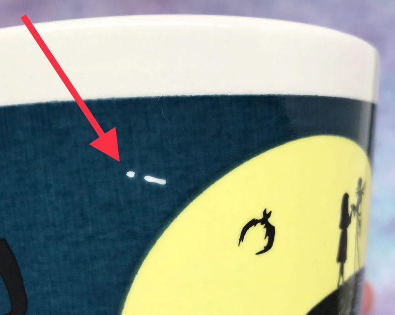May include: A ceramic mug with a dark blue background and a yellow moon design. The mug features black silhouettes of a bat, a figure, and a tree. The top of the mug is white.