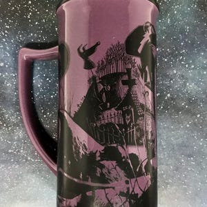 May include: Purple ceramic travel mug with a black lid and handle. The mug features a black graphic design of a spooky graveyard scene with characters. The mug is tall and cylindrical, perfect for hot or cold beverages.