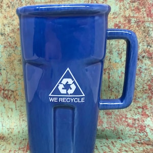 May include: A glossy blue ceramic mug shaped like a recycling bin with a square handle. The front features a white triangular recycling symbol above the text WE RECYCLE. The mug has a vertical molded design mimicking the ridges of a bin.
