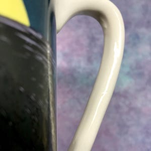 May include: Close-up of a white ceramic mug handle. The handle is curved and smooth, with a glossy finish. The mug's body features a black and yellow design. The background is a blurred mix of purple and blue.