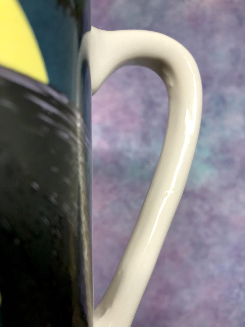 May include: Close-up of a white ceramic mug handle. The handle is curved and smooth, with a glossy finish. The mug's body features a black and yellow design. The background is a blurred mix of purple and blue.