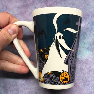 May include: White ceramic mug featuring Zero from The Nightmare Before Christmas. The mug has a dark blue background with images of pumpkins, bats, and a tombstone. The handle is white, and the mug is held by a hand.