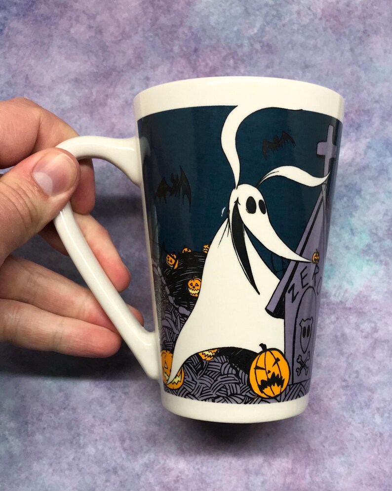 May include: White ceramic mug featuring Zero from The Nightmare Before Christmas. The mug has a dark blue background with images of pumpkins, bats, and a tombstone. The handle is white, and the mug is held by a hand.