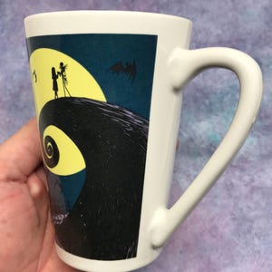 May include: White ceramic mug featuring a design from The Nightmare Before Christmas. The design includes a yellow full moon, silhouettes of Jack and Sally, and a bat against a dark blue background. The mug has a curved handle.