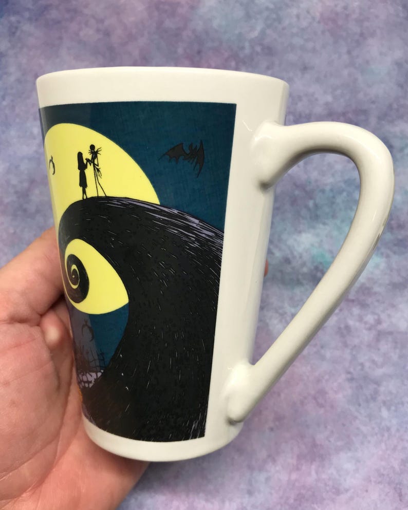 May include: White ceramic mug featuring a design from The Nightmare Before Christmas. The design includes a yellow full moon, silhouettes of Jack and Sally, and a bat against a dark blue background. The mug has a curved handle.