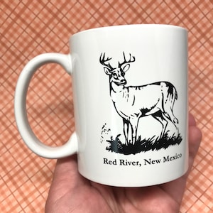 May include: White ceramic mug featuring a black illustration of a deer with antlers. The text "Red River, New Mexico" is printed below the deer. The mug has a curved handle.