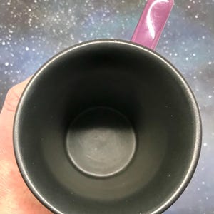 May include: A black ceramic mug with a purple handle. The mug is empty, and the interior is also black. The handle is a glossy, solid purple color. The background is a blurry, speckled pattern of blues, purples, and whites.