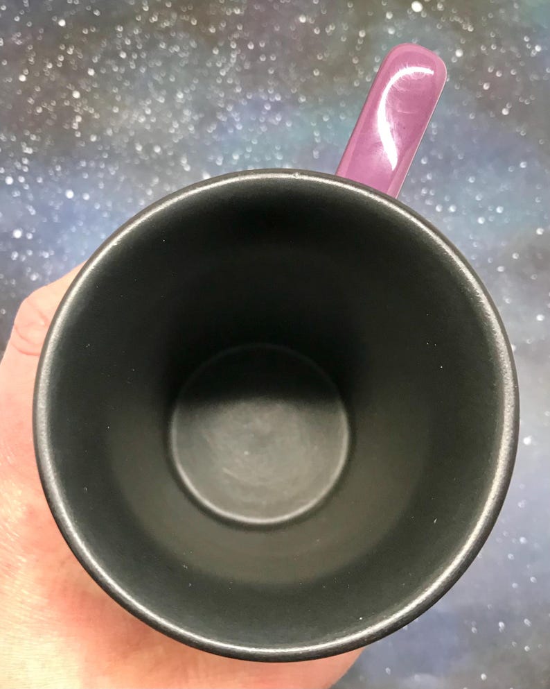 May include: A black ceramic mug with a purple handle. The mug is empty, and the interior is also black. The handle is a glossy, solid purple color. The background is a blurry, speckled pattern of blues, purples, and whites.