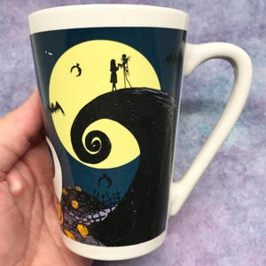 May include: White ceramic mug featuring a design from The Nightmare Before Christmas. The mug has a curved handle and a wraparound illustration of Jack and Sally silhouetted against a full moon, with bats and pumpkins.