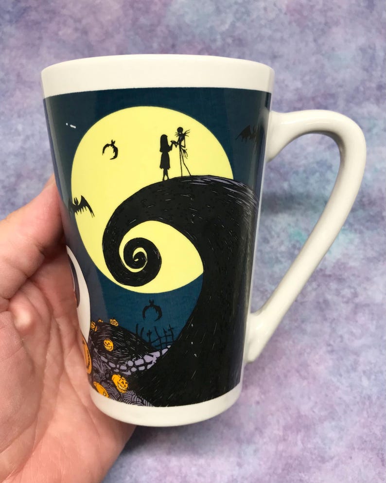 May include: White ceramic mug featuring a design from The Nightmare Before Christmas. The mug has a curved handle and a wraparound illustration of Jack and Sally silhouetted against a full moon, with bats and pumpkins.