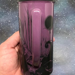 May include: Purple enamel mug with a black rim and handle. The mug features a black graphic design of a spooky forest scene with a full moon and a silhouetted figure. The mug is held in a hand.