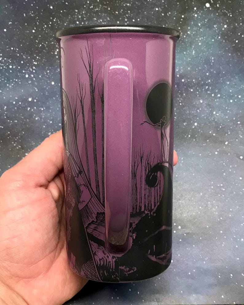 May include: Purple enamel mug with a black rim and handle. The mug features a black graphic design of a spooky forest scene with a full moon and a silhouetted figure. The mug is held in a hand.