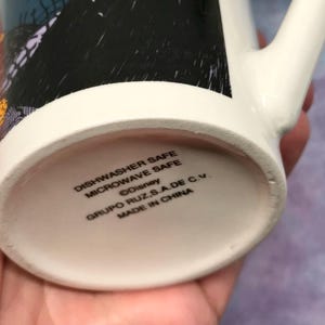May include: White ceramic mug with a black and orange design. The bottom of the mug has the text "Dishwasher Safe Microwave Safe &copy;Disney GRUPO RUZ S.A.DE C.V. MADE IN CHINA". The mug has a white handle.