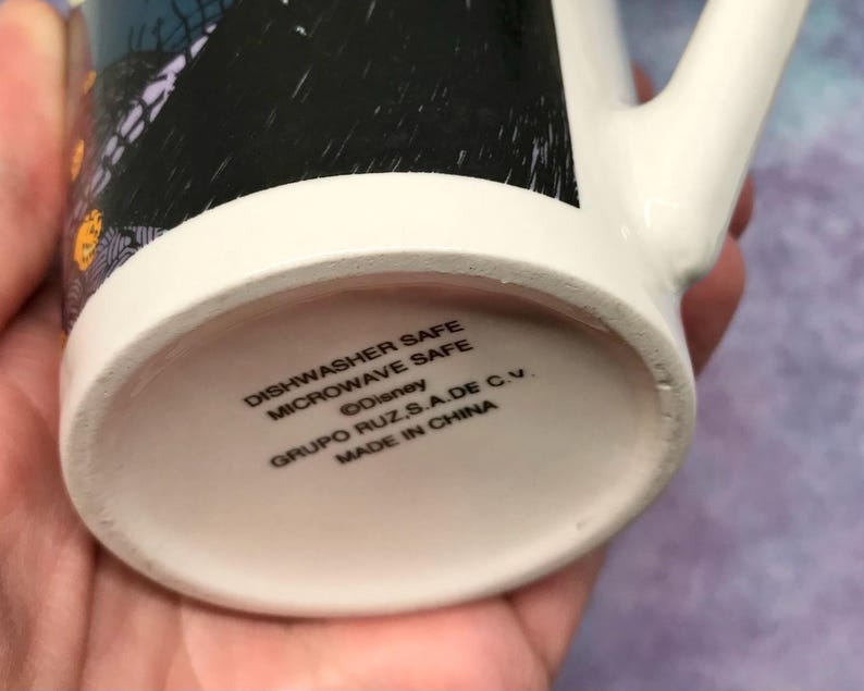 May include: White ceramic mug with a black and orange design. The bottom of the mug has the text "Dishwasher Safe Microwave Safe &copy;Disney GRUPO RUZ S.A.DE C.V. MADE IN CHINA". The mug has a white handle.