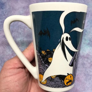 May include: White ceramic mug featuring a Halloween-themed design. The mug showcases a cartoon ghost character with a long, flowing white sheet, bats, and jack-o'-lanterns against a dark blue background. The mug has a curved handle.