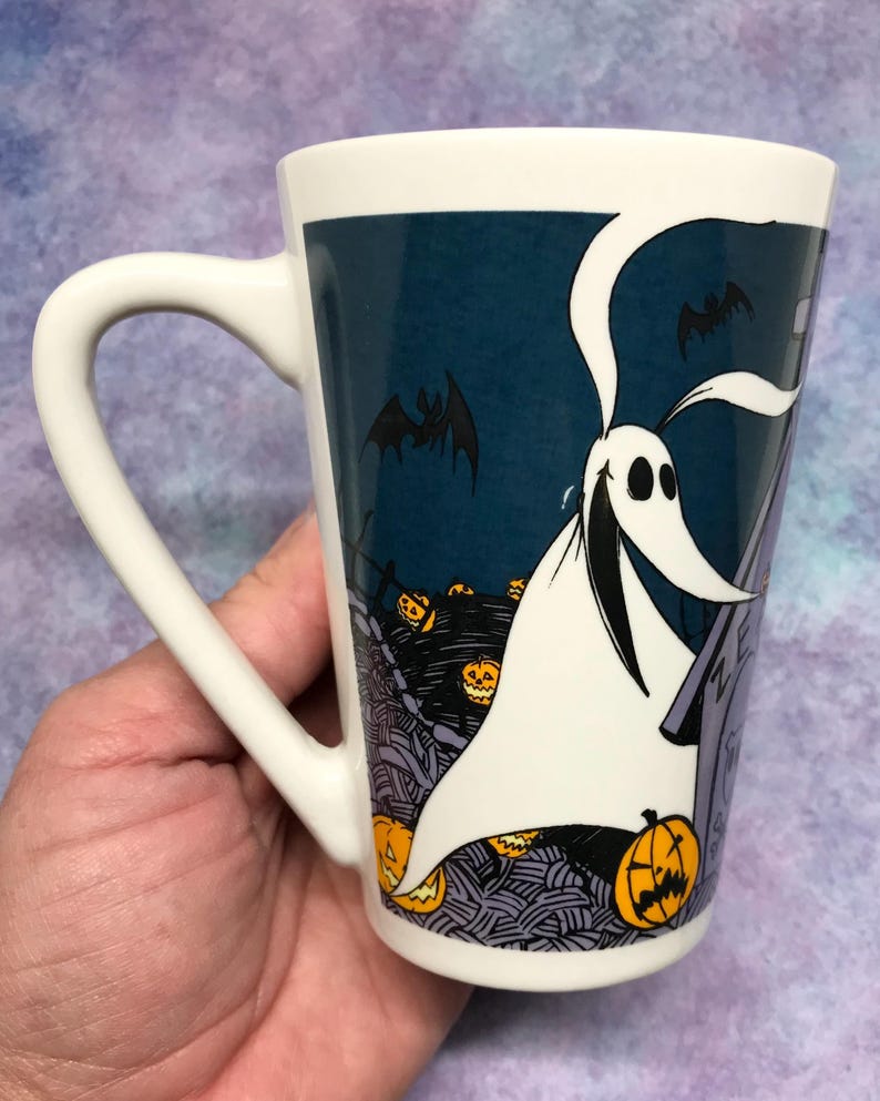 May include: White ceramic mug featuring a Halloween-themed design. The mug showcases a cartoon ghost character with a long, flowing white sheet, bats, and jack-o'-lanterns against a dark blue background. The mug has a curved handle.