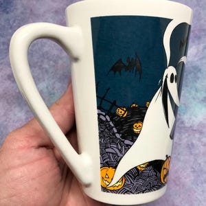 May include: White ceramic Halloween mug with a wraith design. The mug features a wraith with a bat and jack-o'-lanterns against a dark blue background. The mug has a curved handle.
