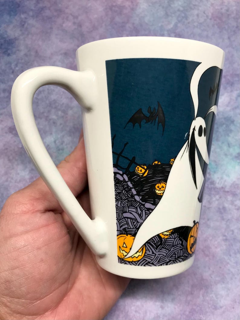 May include: White ceramic Halloween mug with a wraith design. The mug features a wraith with a bat and jack-o'-lanterns against a dark blue background. The mug has a curved handle.