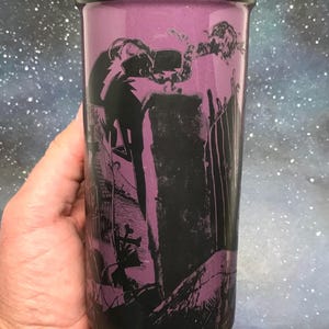 May include: A tall, purple tumbler with a black rim and a spooky graveyard scene printed on the side. The design features a silhouetted figure, tombstones, and a fence. The background is a starry night sky.