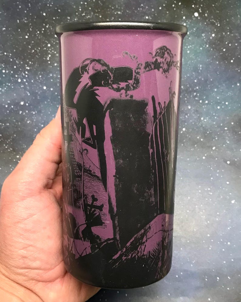 May include: A tall, purple tumbler with a black rim and a spooky graveyard scene printed on the side. The design features a silhouetted figure, tombstones, and a fence. The background is a starry night sky.