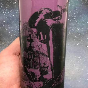 May include: A tall, black and purple tumbler featuring a spooky graveyard scene with a figure and the word "ZERO". The tumbler has a black rim and a purple interior, perfect for cold beverages.