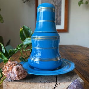 May include: A vibrant blue ceramic tea set, featuring a lidded tea pot and saucer, adorned with gold bands. The tea set is displayed on a wooden surface, with a framed picture in the background. The tea pot is approximately 6 inches tall.