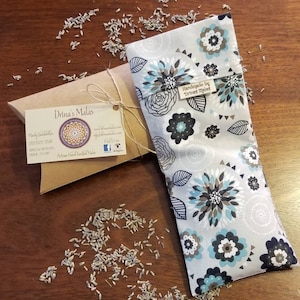 May include: A gray and white floral fabric eye pillow with a brown label that says "Handmade by Drina's Malas". The pillow is filled with lavender and is surrounded by lavender seeds.