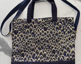 May include: A square tote bag featuring a repeating pattern of dark blue, gold, and light purple feather-like shapes on the upper portion. The bottom section is solid dark blue fabric. It includes two dark blue handles and a long adjustable strap.