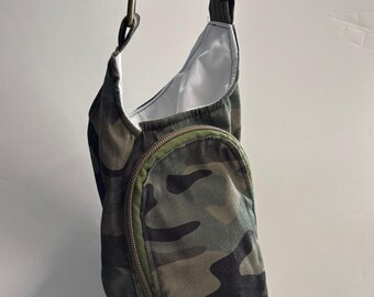 May include: A camouflage print sling bag featuring a vertical zippered front pocket with olive green trim, a white interior lining, and an adjustable black fabric shoulder strap attached with bronze-toned metal hardware rings.