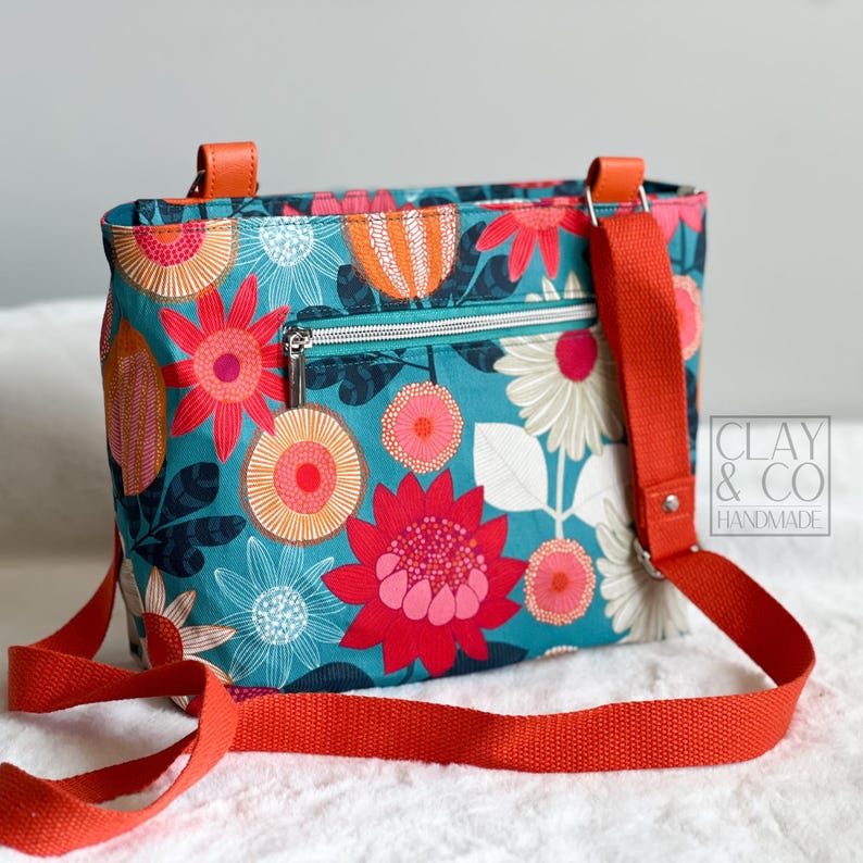A floral crossbody bag with a teal background and vibrant red, orange, and pink flower patterns. The fabric bag has an orange strap and handles, a silver zip, and the text "CLAY & CO HANDMADE". The bag was made using the Phillippa Crossbody sewing pattern.