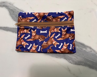May include: A rectangular zippered pouch featuring a repeating pattern of brown dogs and white bones on a dark blue background. The pouch has a tan zipper closure running horizontally across the top section, providing a secure opening for the bag.