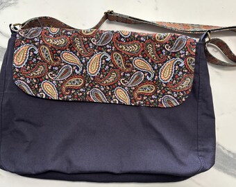 May include: A navy blue messenger bag with a flap featuring a paisley pattern in red, yellow, and blue. The bag has a matching patterned strap and a solid navy blue body. The bag is designed for carrying items.