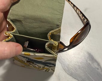 May include: A vertical fabric pouch with a sage green interior and a flap closure featuring a gold-tone snap button. The exterior fabric displays a botanical pattern with yellow and dark blue leaves. A pair of brown tortoiseshell sunglasses rests nearby.
