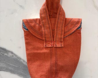 May include: A textured orange fabric bag featuring a single integrated handle at the top. The bag has a structured, tapered shape with visible dark vertical stitching lines running down the center and along the handle, and a black base.