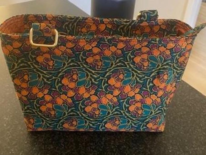 A rectangular tote bag featuring a floral design in teal, orange, and purple. The bag has two handles with gold-coloured metal hardware. The interior lining is a solid orange colour. The bag was made using the Phillippa Crossbody sewing pattern.