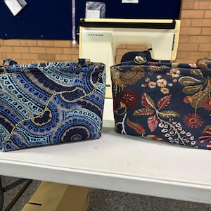 Two fabric tote bags with handles on a white table. One has a blue and gold patterned design, the other a floral pattern in navy, red, and gold. A sewing machine is in the background. The bags were both made using the Phillippa Crossbody sewing pattern.