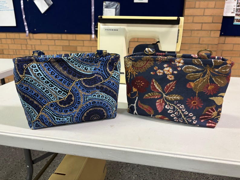 Two fabric tote bags with handles on a white table. One has a blue and gold patterned design, the other a floral pattern in navy, red, and gold. A sewing machine is in the background. The bags were both made using the Phillippa Crossbody sewing pattern.