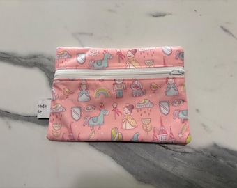 May include: A rectangular pink zippered pouch featuring a repeating pattern of cartoon figures including princesses, unicorns, rainbows, castles, and wands. A white zipper runs across the top, and a small white fabric tag is attached to the side.