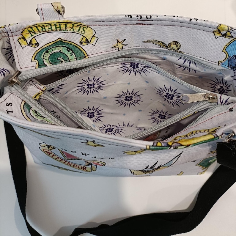 A detail of a light blue tote bag with a black strap, featuring a repeating pattern of Hogwarts crests and symbols. The bag has two zippered compartments and a light-coloured interior with a starburst pattern. The bag has a top handle and was made using the Phillippa Crossbody sewing pattern.