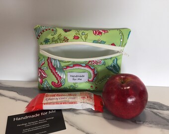 May include: A light green zippered pouch with a colorful floral paisley pattern and a white label reading Handmade for Me. Next to the pouch is a red apple and a Great Grains Chewy Choc Chip snack bar. A black business card with white text is also visible.