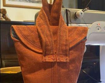 May include: An orange tote bag with a long handle. The bag is made of a textured fabric and has a flap closure. The bag is sitting on a white surface.