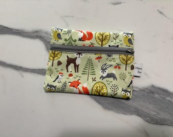 May include: A rectangular fabric zipper pouch featuring a light green background with a repeating pattern of woodland animals including foxes, owls, deer, and rabbits, alongside trees and mushrooms. A gray zipper runs across the top of the pouch.