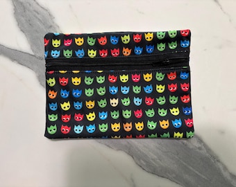 May include: A rectangular black zippered pouch featuring a repeating pattern of small, stylized cartoon faces in vibrant colors including red, yellow, blue, orange, and green. The pouch has a black zipper closure along the top edge.