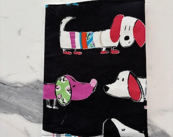 May include: A rectangular fabric book cover featuring a black background with a repeating pattern of stylized dogs. The dogs are depicted in various colors including white, pink, and black, with some wearing colorful striped sweaters or green collars.