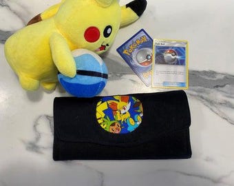 May include: A yellow Pikachu plush toy holding a blue and white plush ball, accompanied by a Pokémon trading card featuring a Poké Ball and a black rectangular wallet with a circular patch displaying colorful cartoon characters.